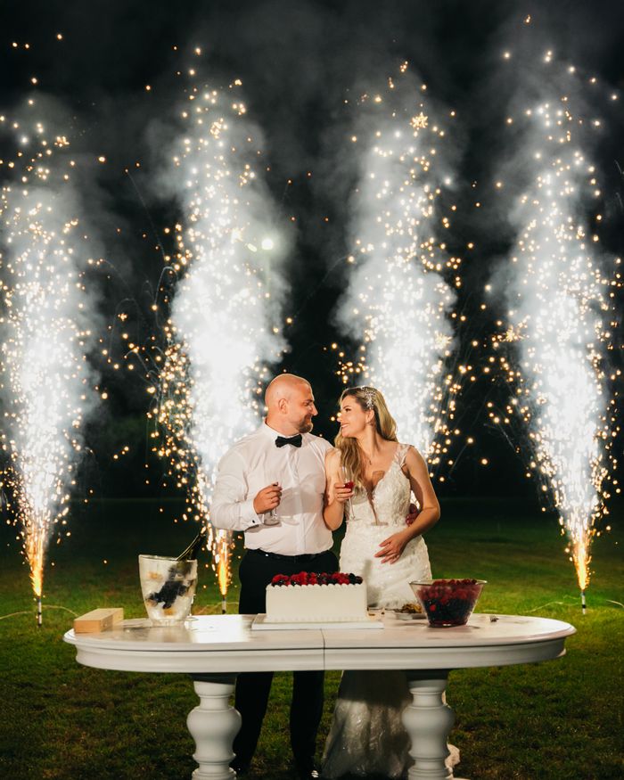 A couple celebrates their wedding with sparklers and a cake cutting ceremony outdoors at night.