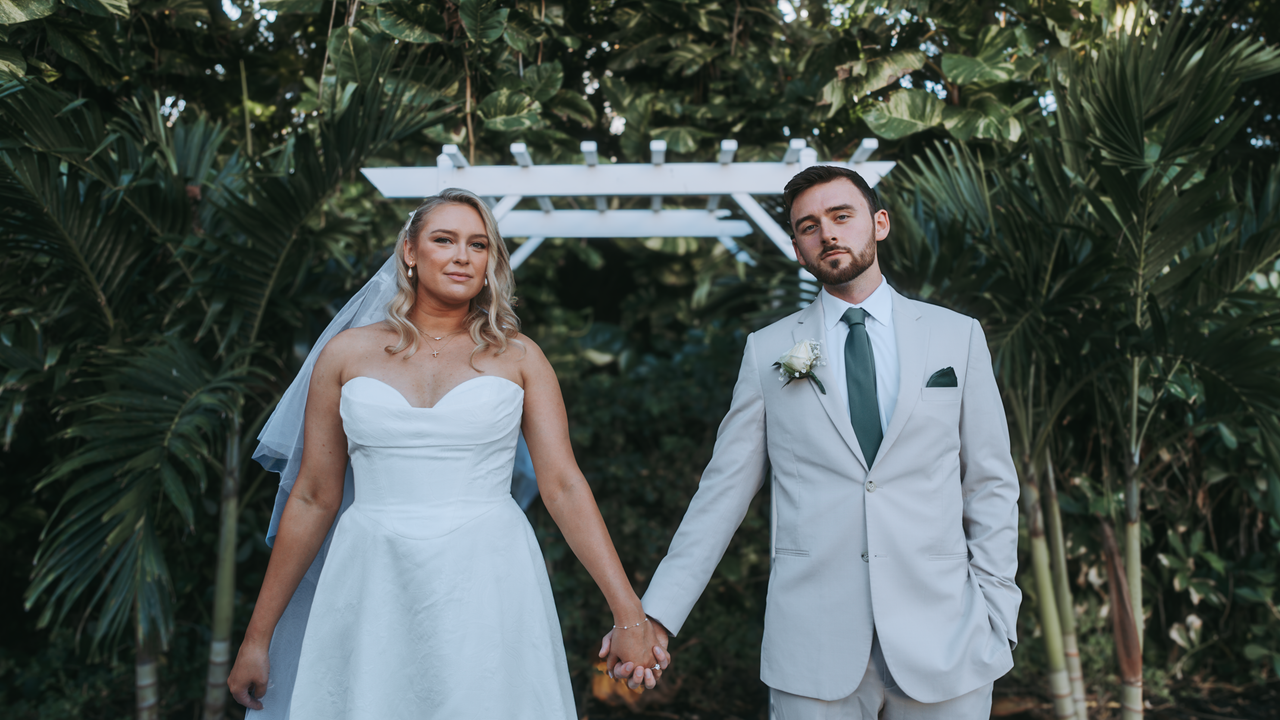A bride and groom holding hands under a white arch in an outdoor wedding setting.